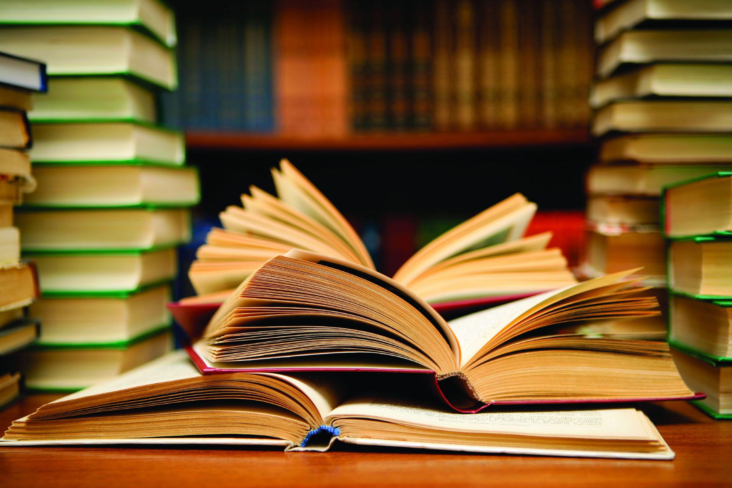 书在桌子上打开, surrounded by other books stacked on the same table and in a bookshelf behind the table.