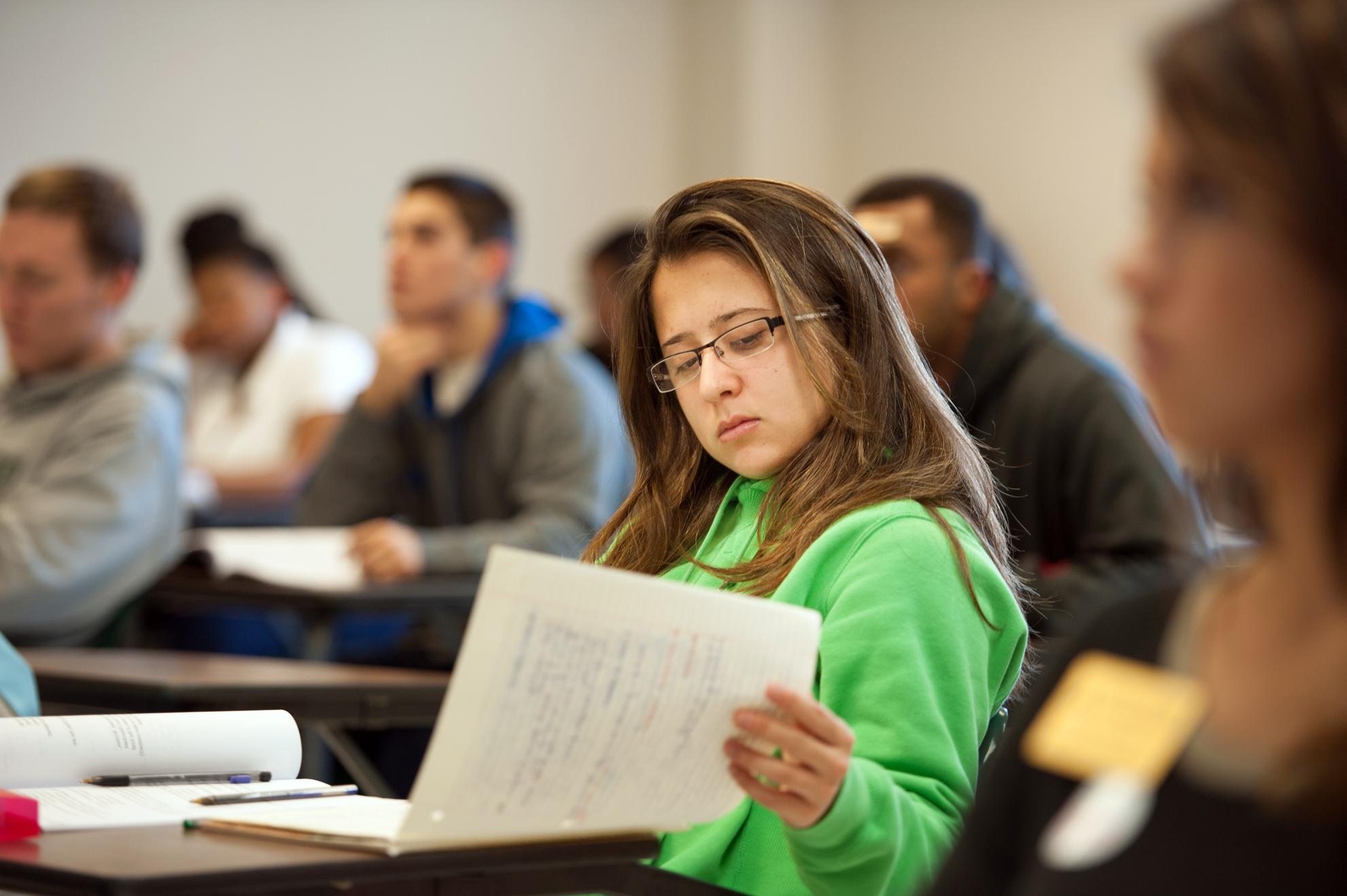 Student sitting at desk in a full classroom, reviewing written notes.