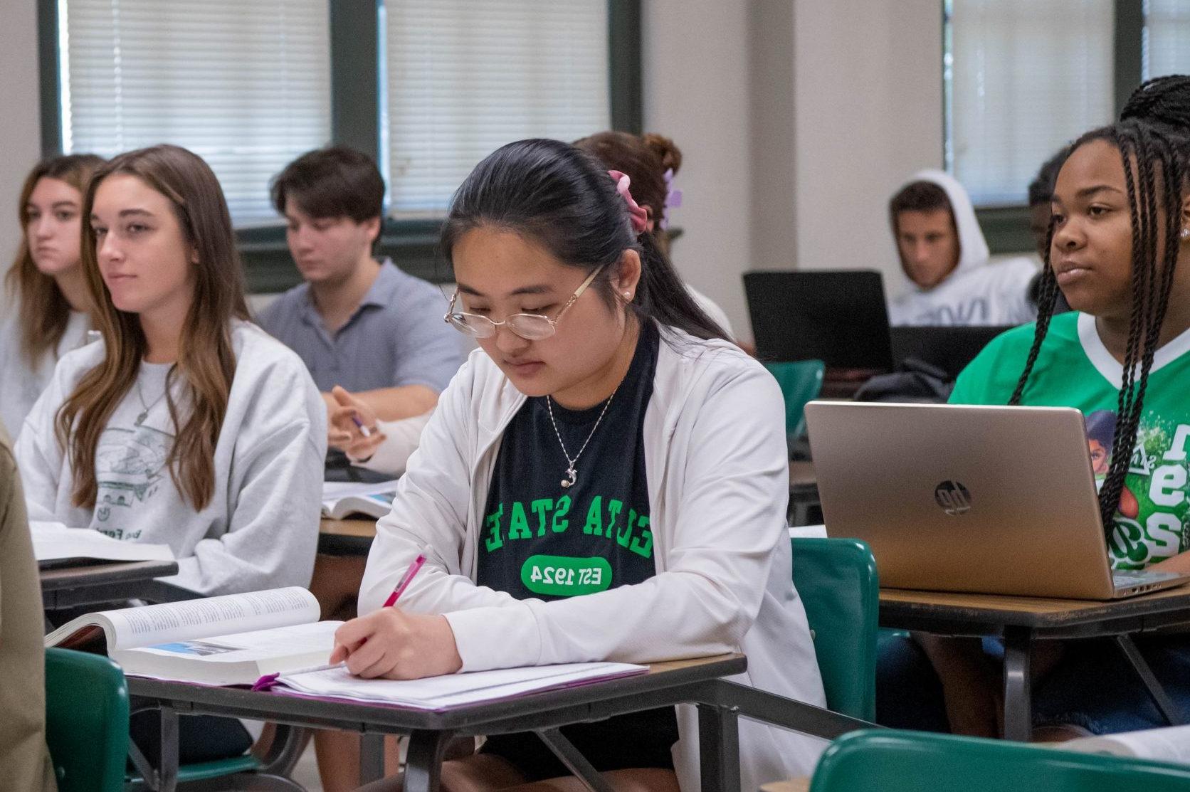 Student sitting at a desk in a full classroom, 在纸上做笔记 with textbook open.