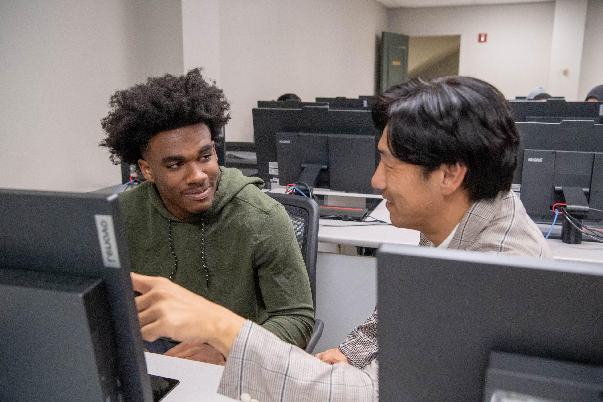 Professor sitting next to student in a computer lab, while pointing at students computer screen.